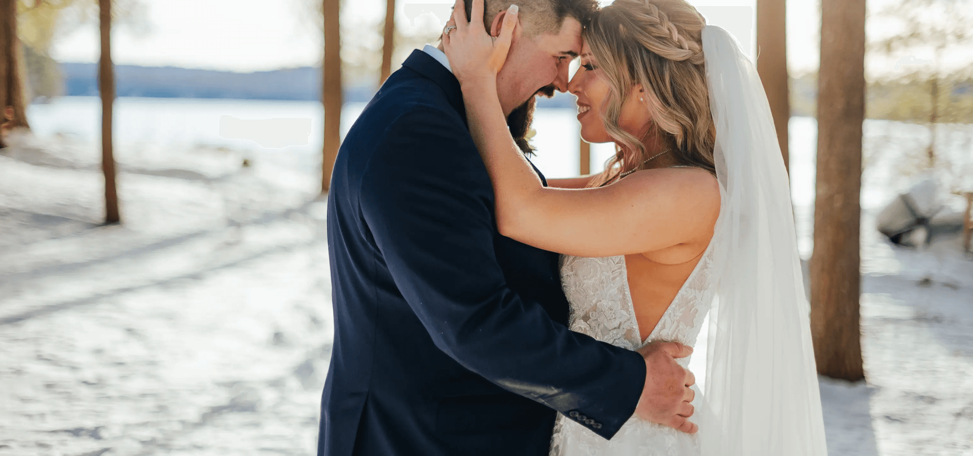 A couple embraces warmly in a snowy outdoor setting, surrounded by trees and a serene lake backdrop.