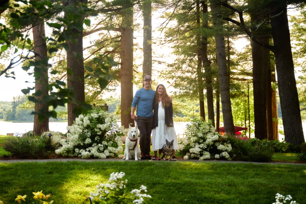 A smiling couple stands with their two dogs in a lush, green garden by a lake.