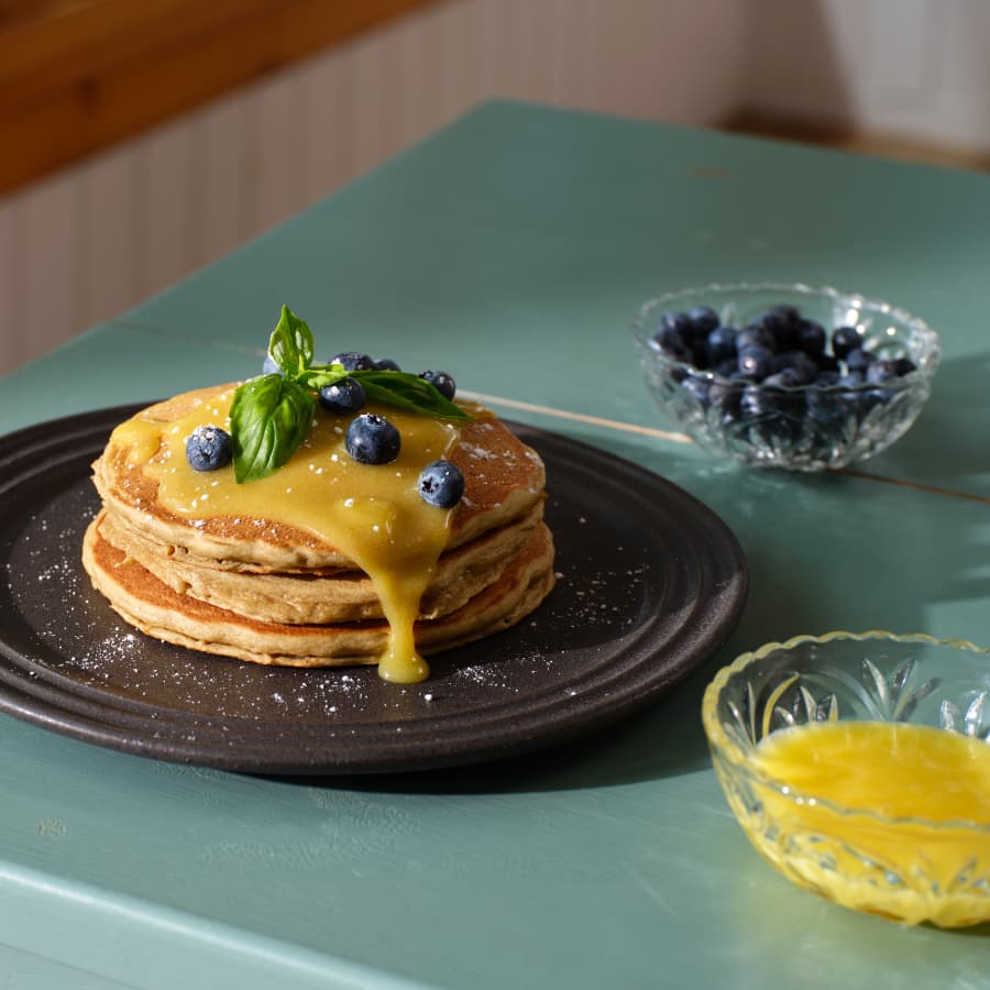 Stack of pancakes topped with a citrus sauce, blueberries, and fresh basil, served with a bowl of blueberries on the side.