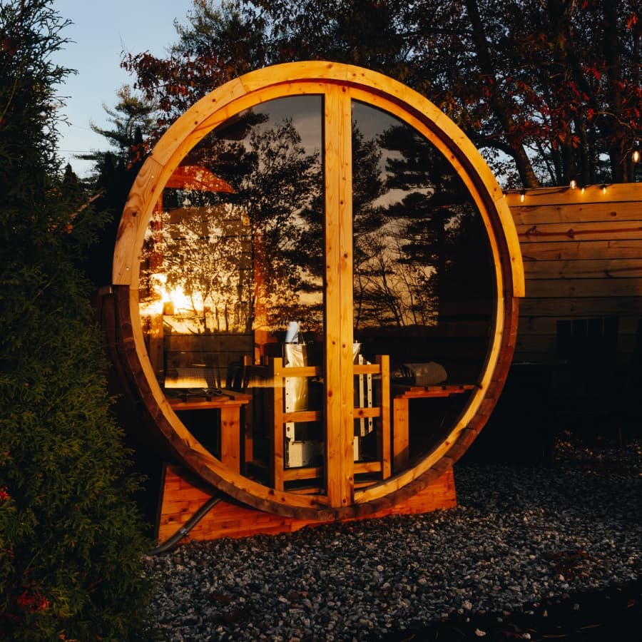 A circular wooden structure with large glass panels reflecting a sunset amidst trees.