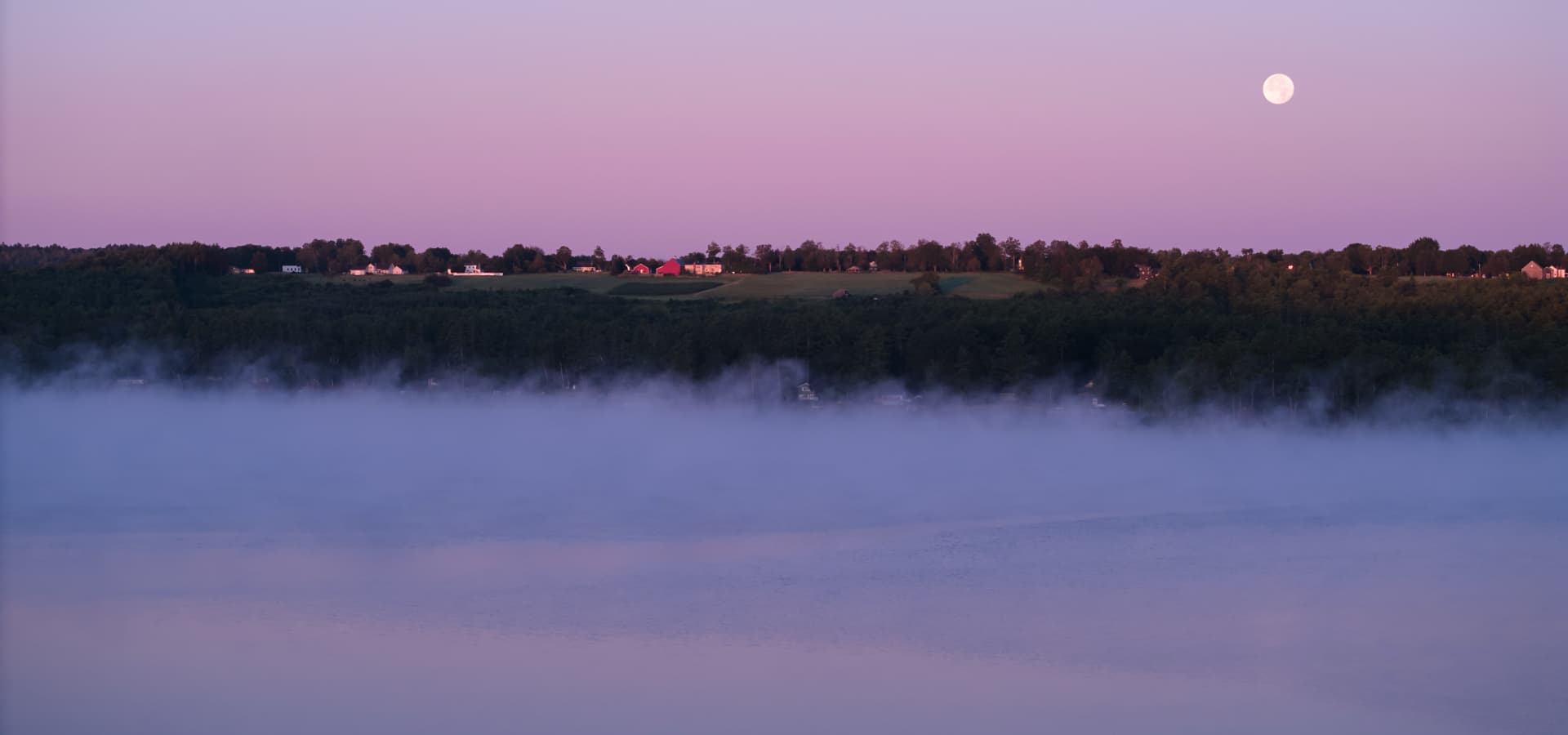 A tranquil landscape at dusk featuring a full moon above misty waters and a distant tree-lined horizon with scattered homes.