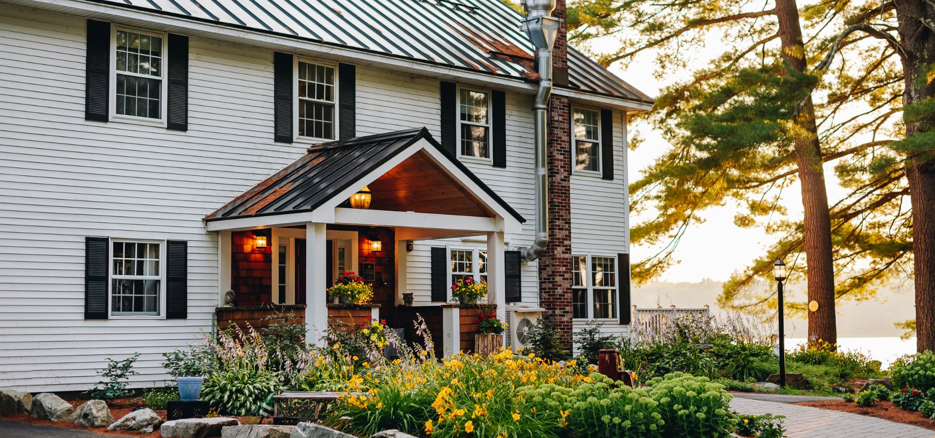 A cozy white house with a brown shingle porch is surrounded by colorful flowers and trees.