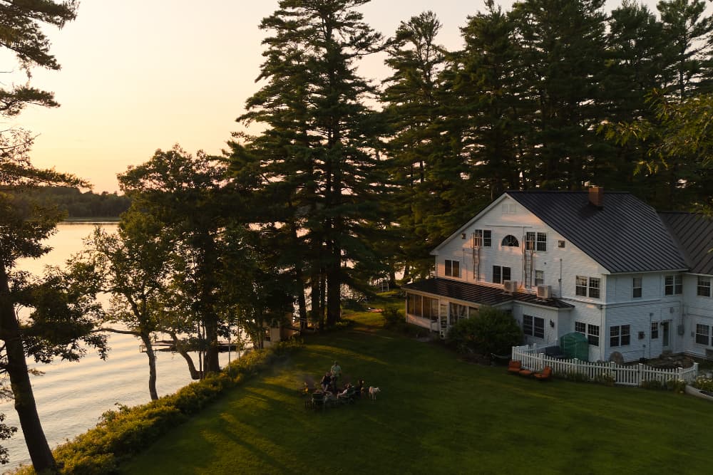A lakeside house surrounded by trees at sunset, with a gathering of people on the lawn.