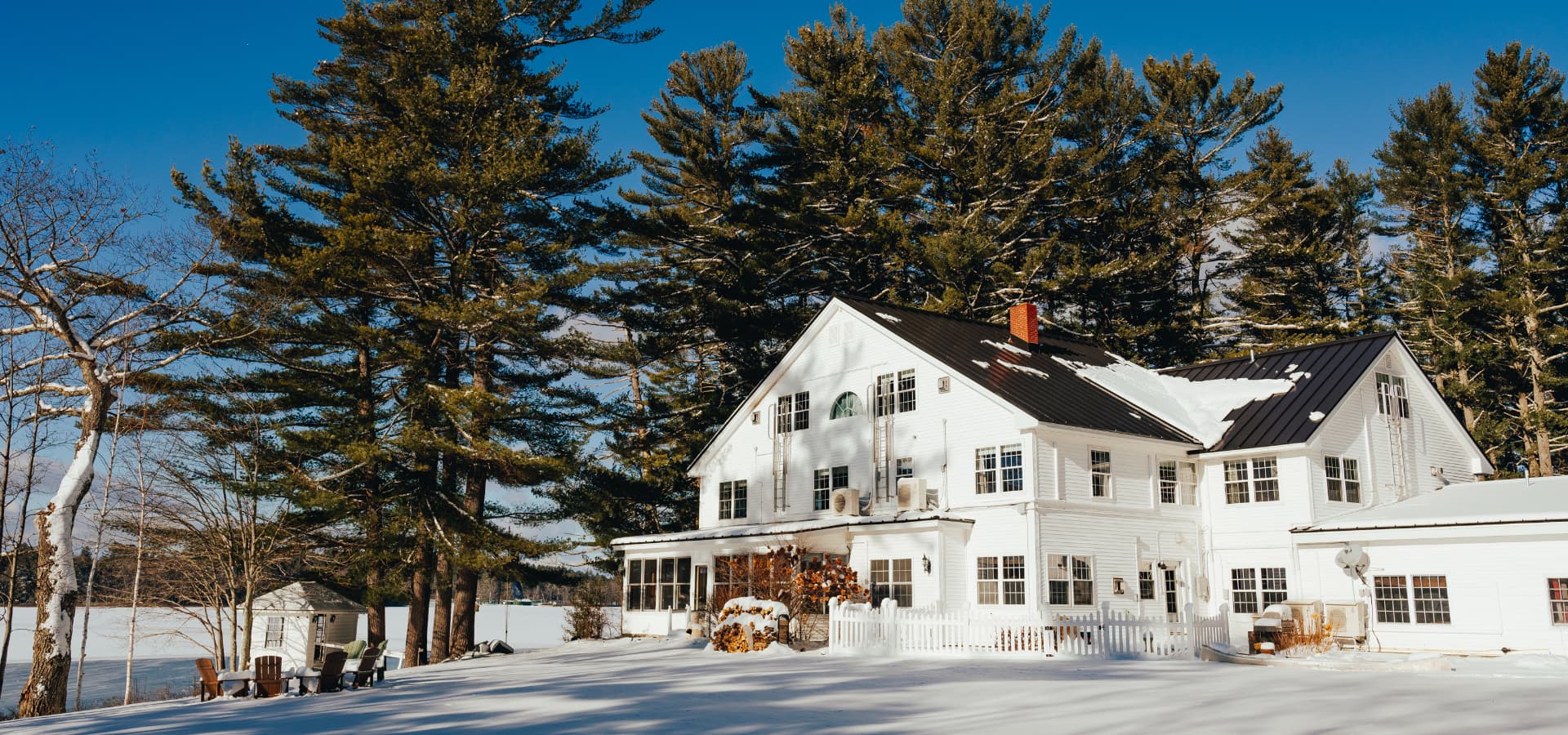 A charming white house surrounded by tall pine trees, set against a clear blue sky and a snowy landscape.