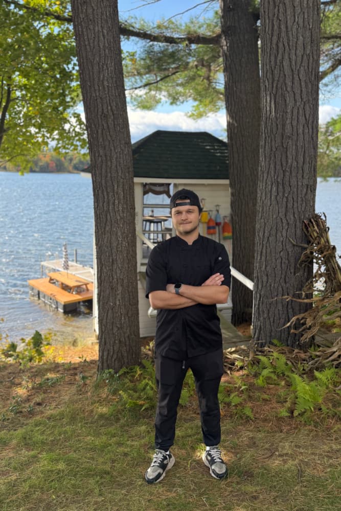 A chef stands by a serene lake in front of a small cabin, framed by tall trees.