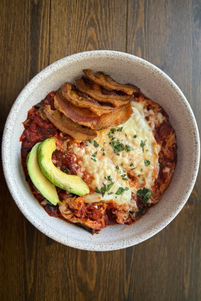 A bowl of cheesy casserole topped with crispy bacon and avocado slices, garnished with herbs.